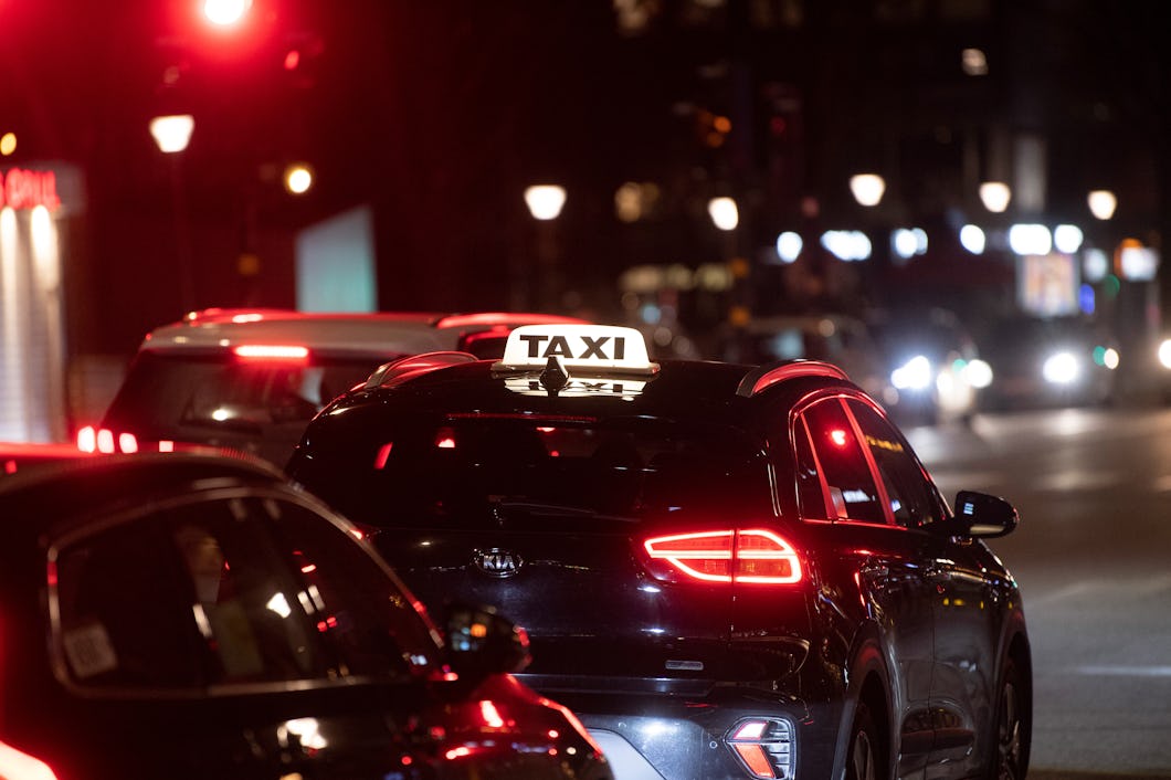 En taxi med ett upplyst takskylt väntar vid ett rödljus på en stadsgata under natten, omgiven av andra fordon och gatlyktor.