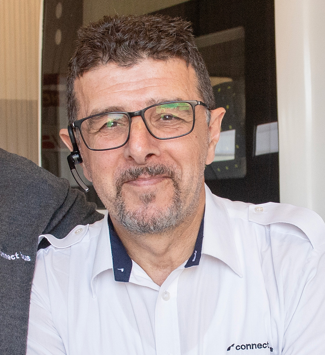 En man med glasögon och headset, klädd i en vit skjorta, tittar mot kameran och ler svagt.