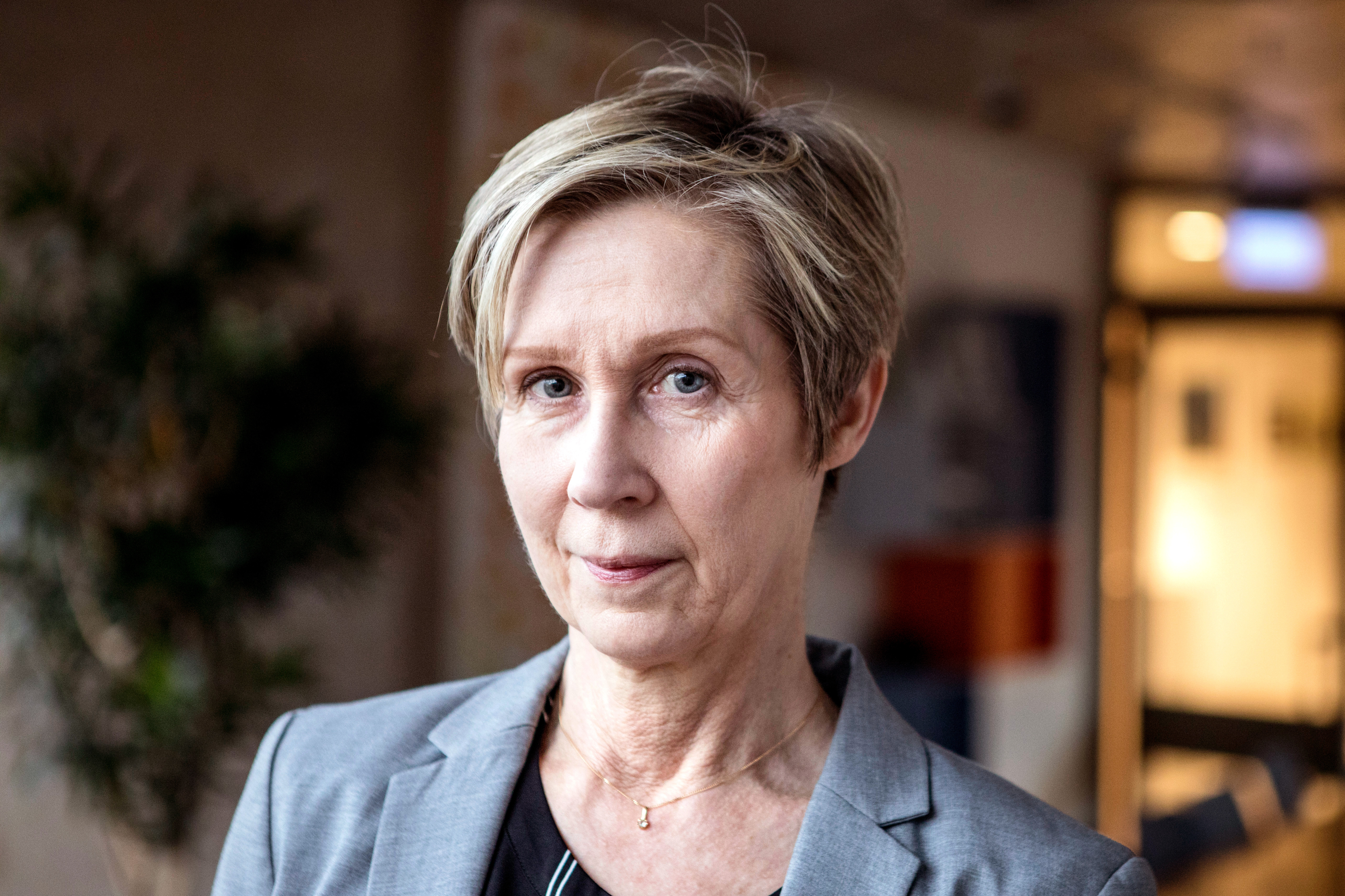 En kvinna i medelåldern med kort blont hår och grå kavaj står inomhus och tittar rakt mot kameran med neutral min.