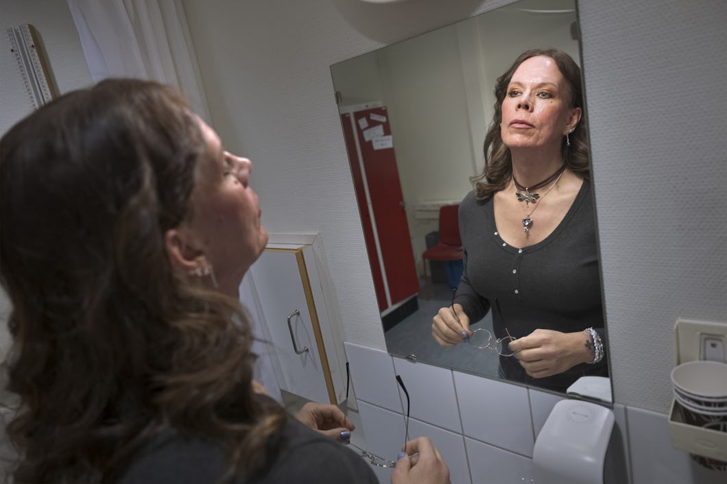 Jackie med långt brunt hår tittar på sig själv i en badrumsspegel. Hon håller glasögon i händerna och har på sig en mörk överdel samt smycken.
