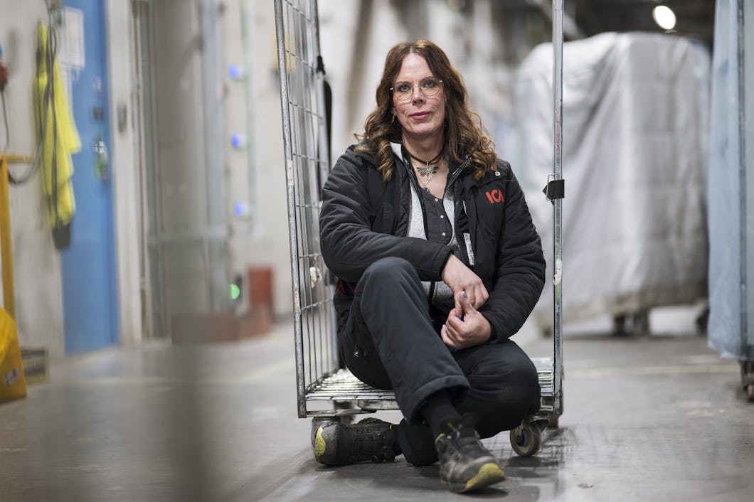 Jackie med långt hår och glasögon sitter på golvet framför en metallvagn i en lager- eller industrimiljö. Personen har på sig jacka, byxor och stövlar i svart färg.