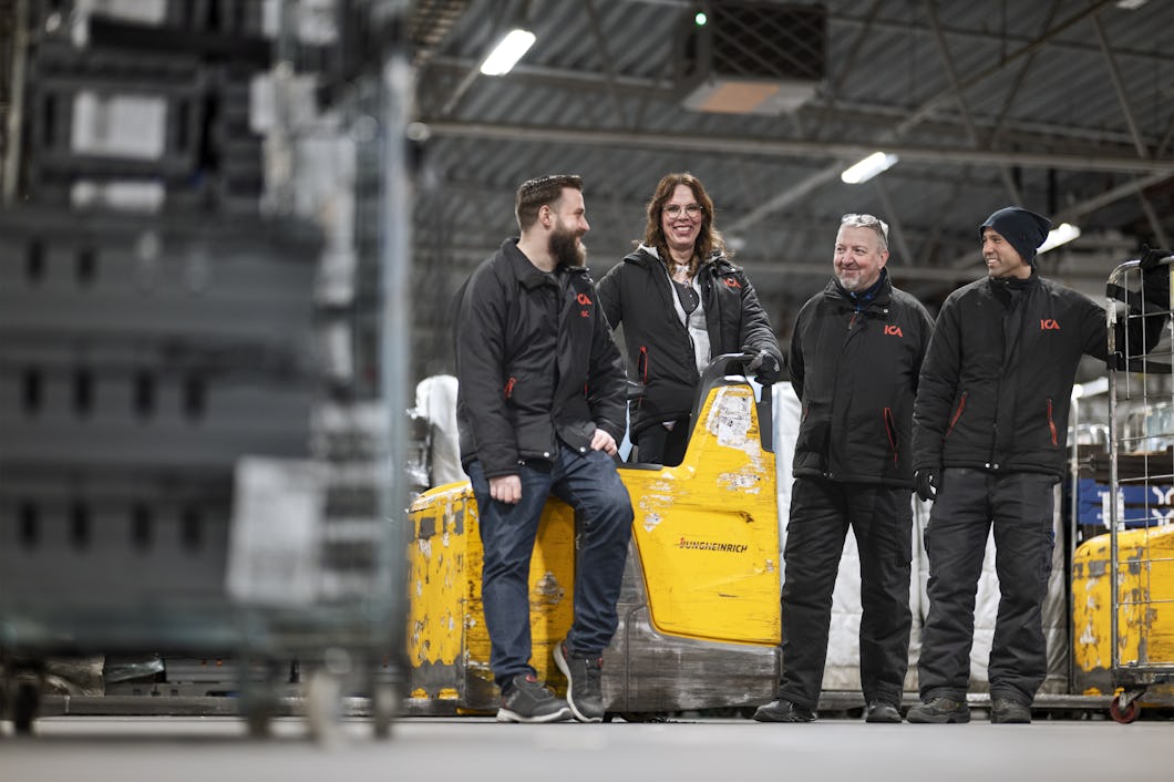 Jackie och tre kollegor står tillsammans i ett lager, leende och samtalande nära en gul palldragare. Industriställningar och utrustning syns i bakgrunden.