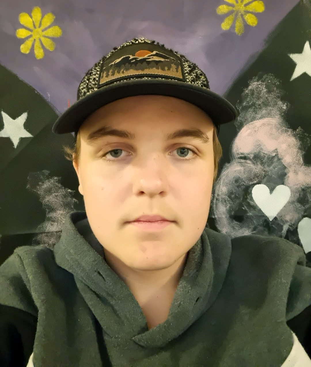 En person med svart hatt och grå huvtröja står vänd mot kameran. Bakom personer syns stjärnor, blommor och ett hjärta på en mörk bakgrund med flera färger.