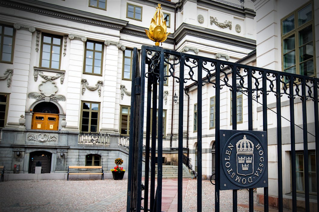 En smidesgrind med en sigill märkt ”Högsta domstolen” står framför en byggnad som tillhör regeringen. Byggnaden har fönster och trappor av sten.
