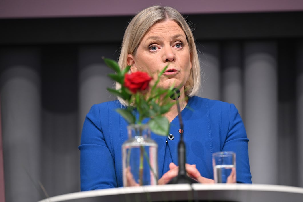 Magdalena Andersson med blont hår och blå kavaj talar vid en talarstol. Framför henne står en röd ros i en glasvas och ett glas med vatten.