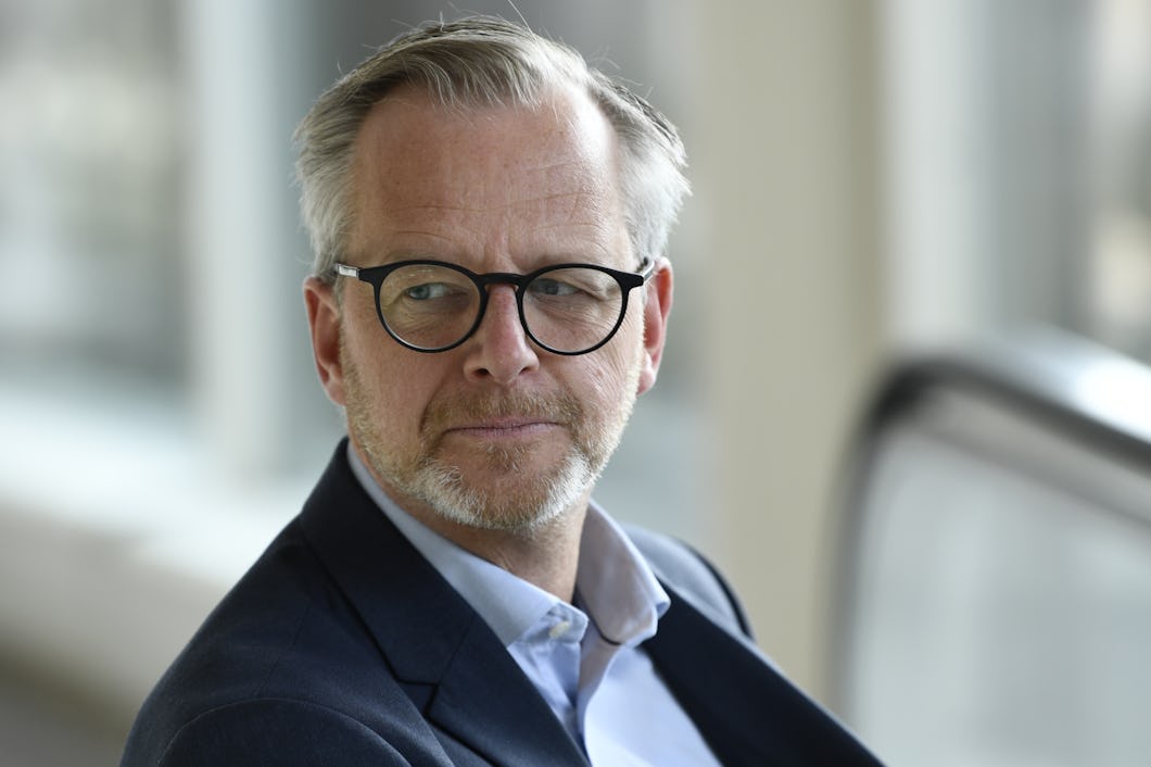En medelålders man med grått hår, skägg och glasögon har på sig en mörkblå kostym och en ljusblå skjorta. Han tittar åt sidan inomhus.