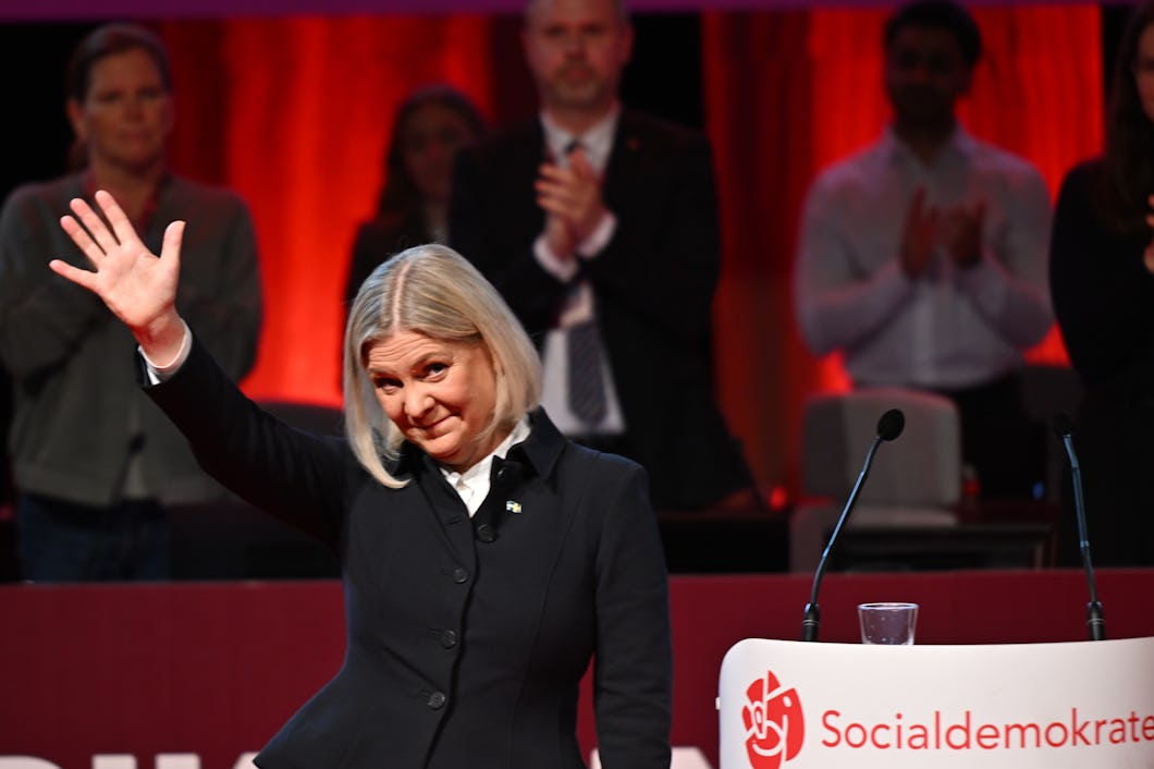 Magdalena Andersson vinkar vid en talarstol med Socialdemokraternas logotyp. Flera personer står och applåderar i bakgrunden.