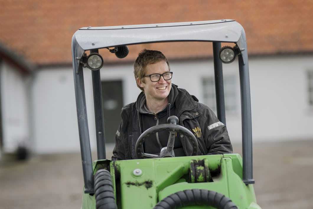 En man med glasögon och svart jacka sitter på ett grönt arbetsfordon. Han ler och i bakgrunden syns en suddig byggnad.