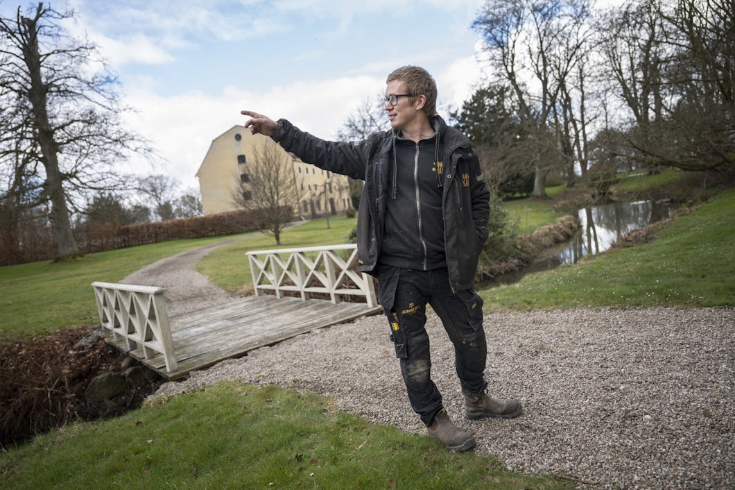 En person iförd arbetskläder står på en grusgång nära en liten bro och pekar åt ett håll, med träd och en byggnad i bakgrunden.