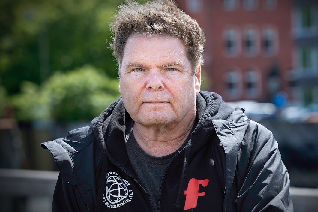Man med kort mörkt hår står utomhus. Han har svart jacka på sig. I bakgrunden syns byggnader och växtlighet.
