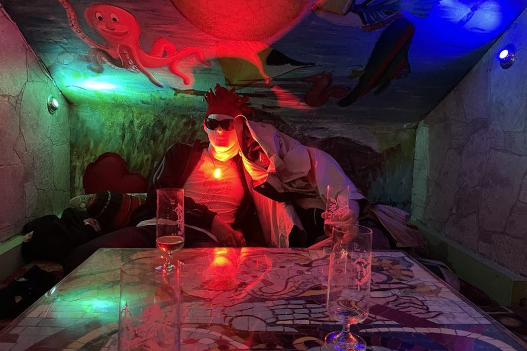 En person med solglasögon och ett rött lysande halsband sitter vid ett mosaikbord i ett rum med väggmålningar och tomma glas.