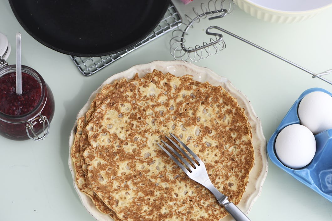 En tallrik med tunna pannkakor har en gaffel ovanpå. Runt tallriken finns ägg, en burk sylt, ett vispverktyg och ett avsvalningsgaller med en stekpanna.