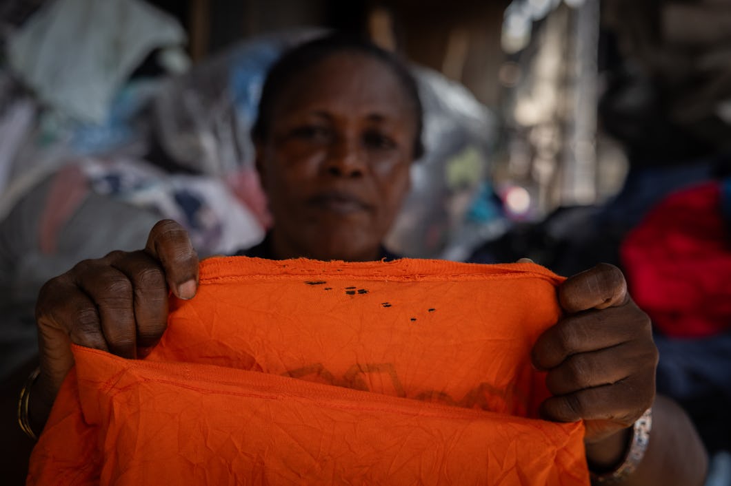 En person håller upp ett orange plagg som har små hål nära kraften, medan kläder och textilier syns suddigt i bakgrunden.