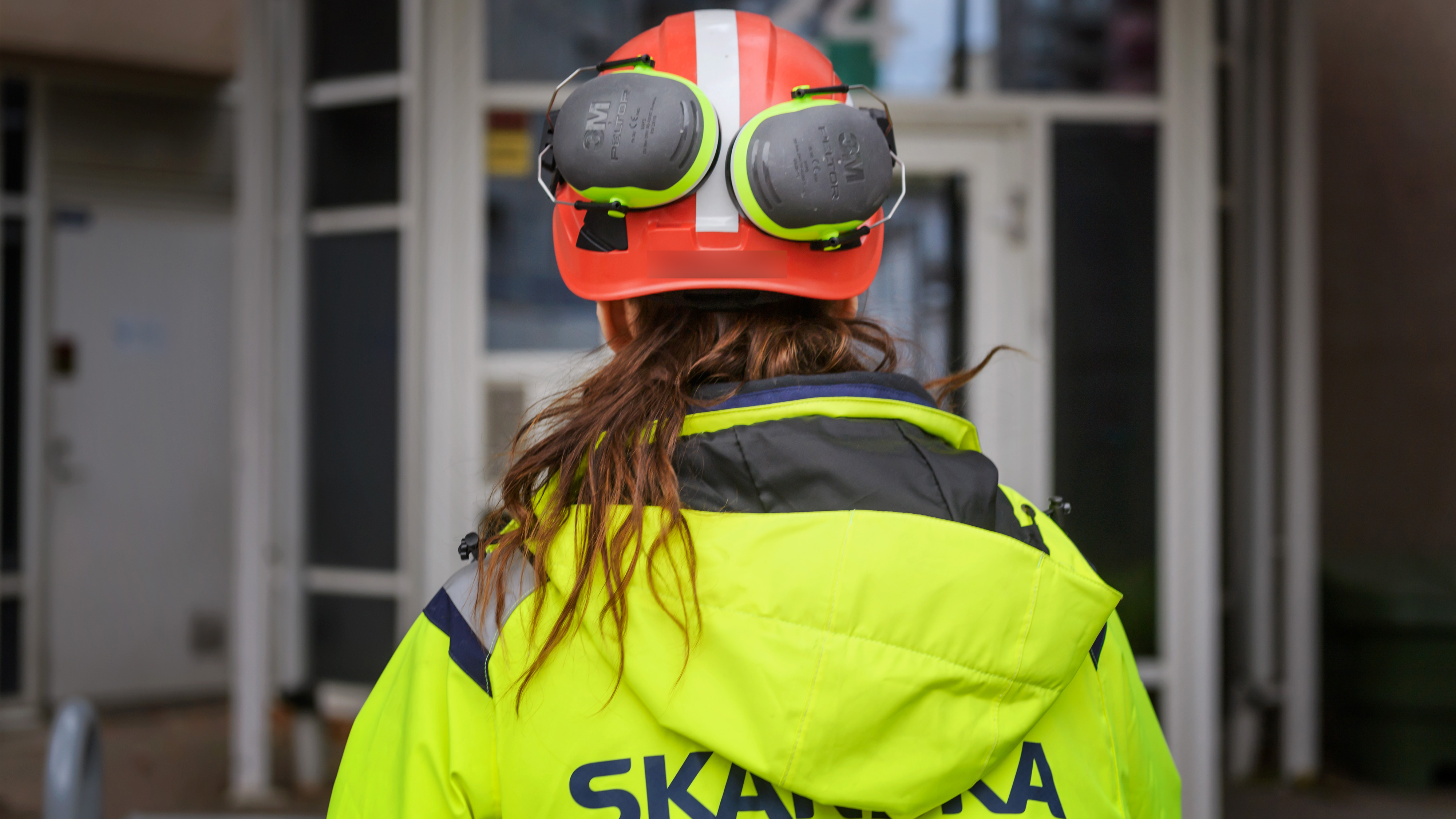 En byggarbetare med varseljacka och röd hjälm med hörselskydd står utanför en byggnad, vänd bort från kameran.