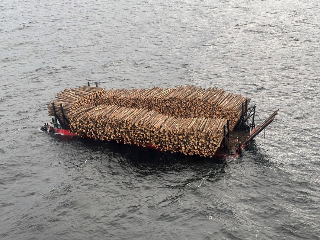 En pråm med timmer lastad i staplar flyter på vatten och omges av vågor, sedd uppifrån.