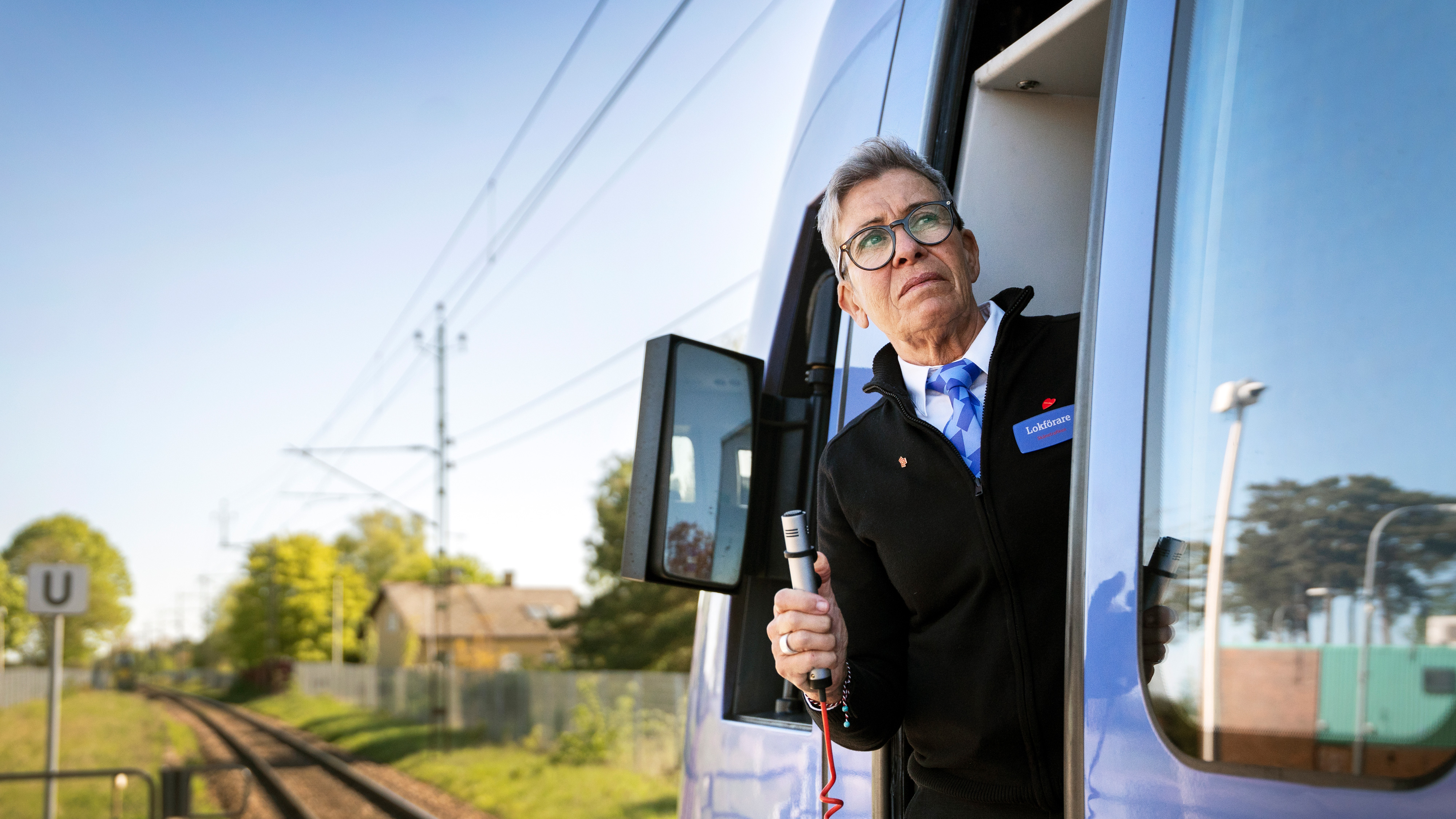 En tågförare med glasögon och namnbricka ser ut genom fönstret i förarhytten. Hen håller i en mikrofon. Utanför syns järnvägsspår och hus.