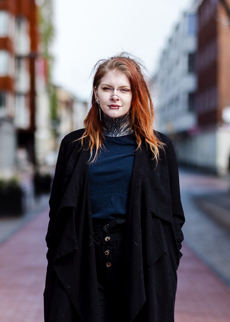 En person med långt rödaktigt hår står på en gågata i en stadsmiljö och har på sig en svart kappa och mörka kläder. Det finns byggnader på båda sidor av gatan.