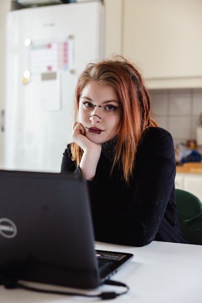 Wildha Eklund med rött hår och piercingar sitter vid ett bord, stödjer hakan mot handen och har en öppen laptop framför sig i ett kök.