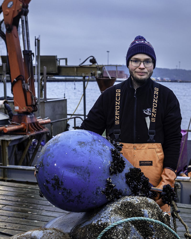 En person med orange overall och blå mössa står på en förtöjd fiskebåt och håller i stora blå bojar som har havsstulpaner på sig.