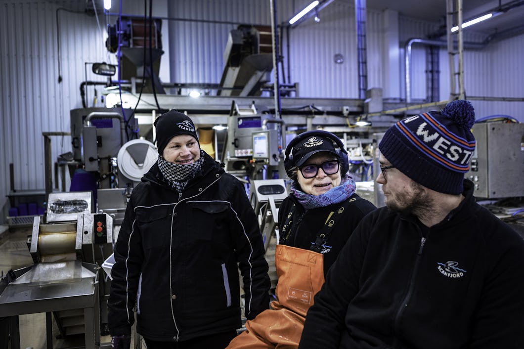 Tre personer med varma kläder och mössor står och sitter i en fabriksmiljö där industriutrustning syns i bakgrunden.