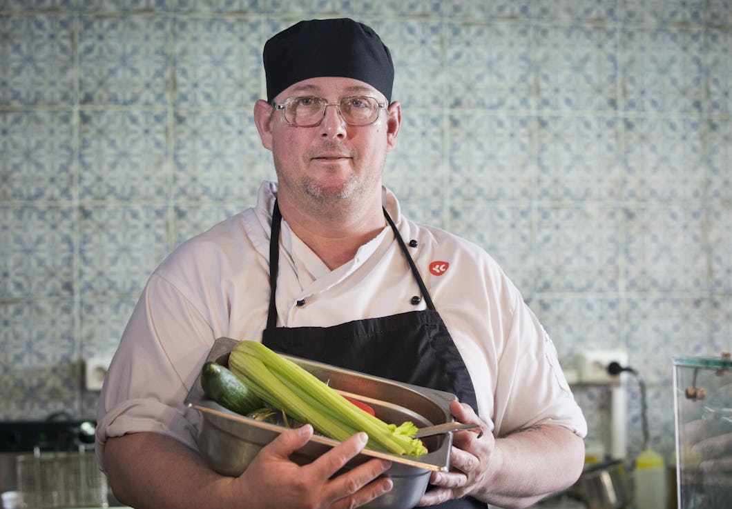 Patrik Nikkinen med arbetskläder och svart mössa håller skålar med grönsaker, bland annat selleri och zucchini, och står i ett kök med väggar av mönstrade kakelplattor.