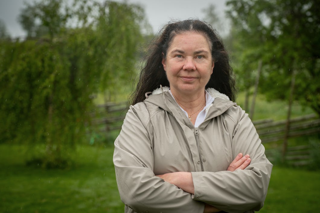 Lena Björk med långt mörkt hår står utomhus med armarna i kors och har på sig en ljus jacka. I bakgrunden syns grönt med träd och gräs.