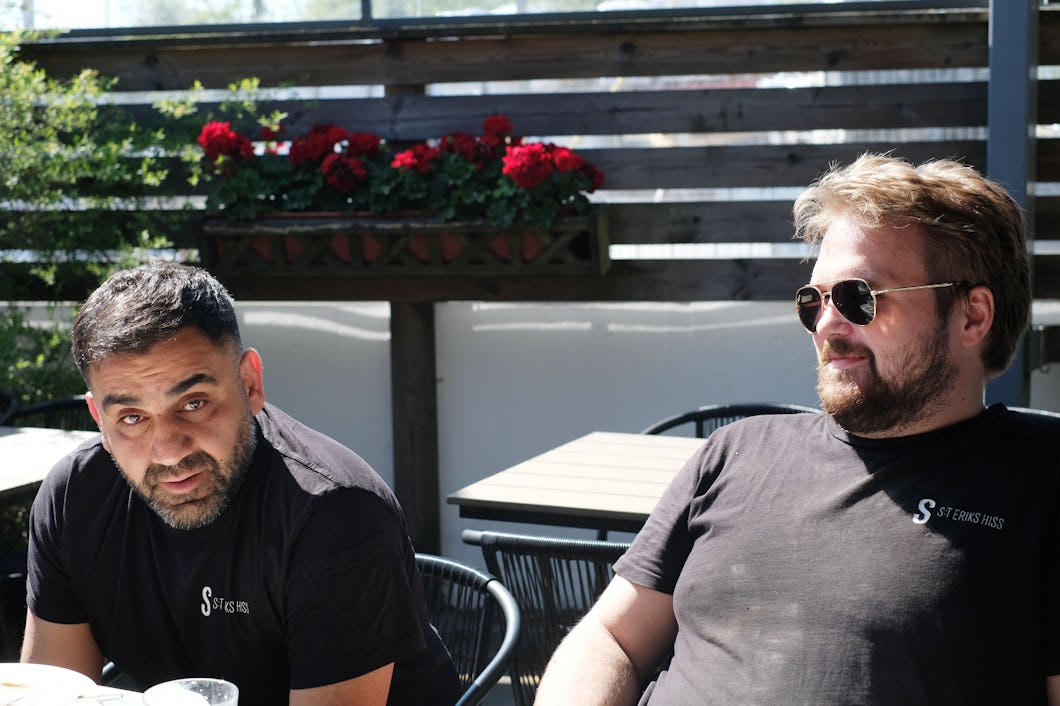 Två män med svarta t-shirts sitter utomhus vid ett bord. Den ena mannen tittar in i kameran, medan den andra, som har solglasögon, tittar åt sidan.