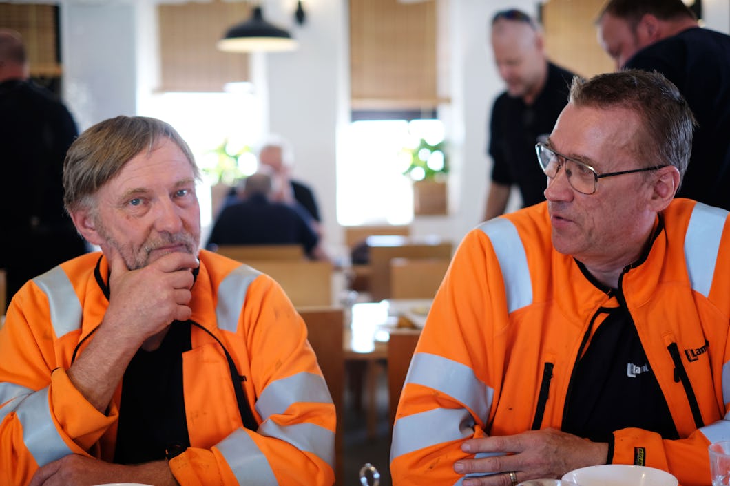 Två män med orangea varseljackor sitter vid ett bord i en matsal och pratar med varandra. Andra personer finns i bakgrunden.