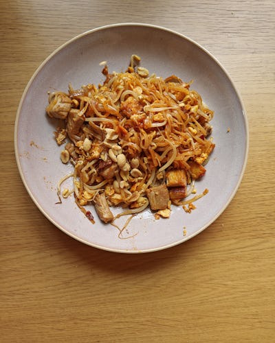 En tallrik pad thai med tofu, böngroddar och krossade jordnötter står på ett bord av trä.