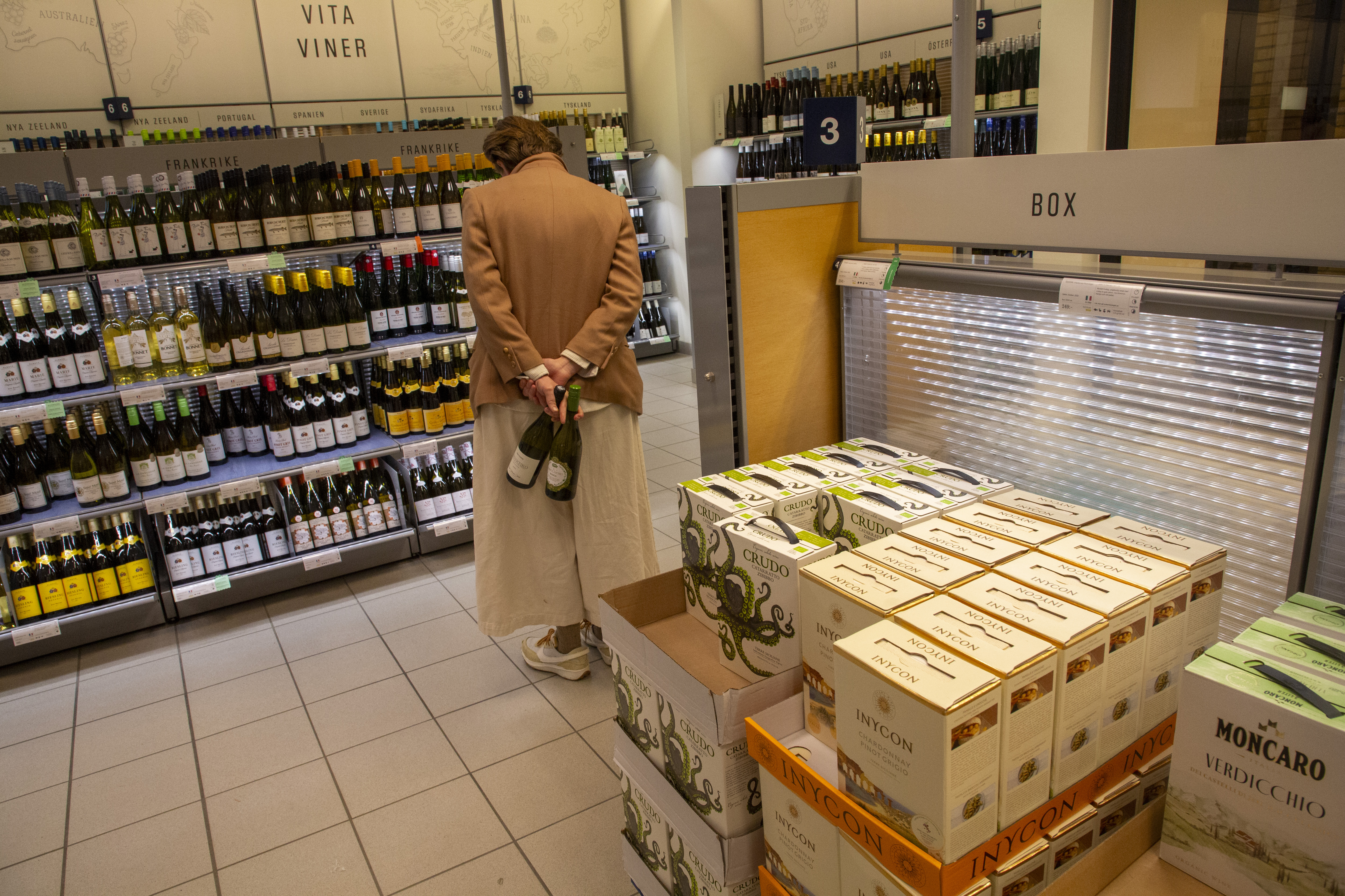 En person med en ljusbrun kappa står i en vinbutiksgång och håller två vinflaskor. Personen är vänd mot hyllor där det finns olika flaskor vin, och bredvid finns lådor med vin.