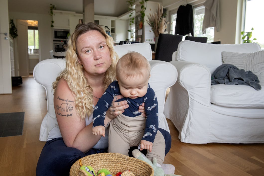 Camilla Dahlgren med långt blont hår sitter på golvet och håller ett spädbarn i vardagsrummet. Där finns vita möbler och leksaker ligger utspridda.