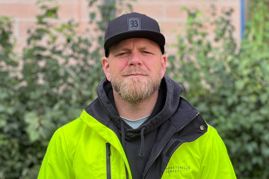 Skyddsombudet Johan Larsson-Hytte med skägg och svart keps har på sig en svart huvtröja och en gul jacka. Han står utomhus utomhus grönska och en tegelvägg.