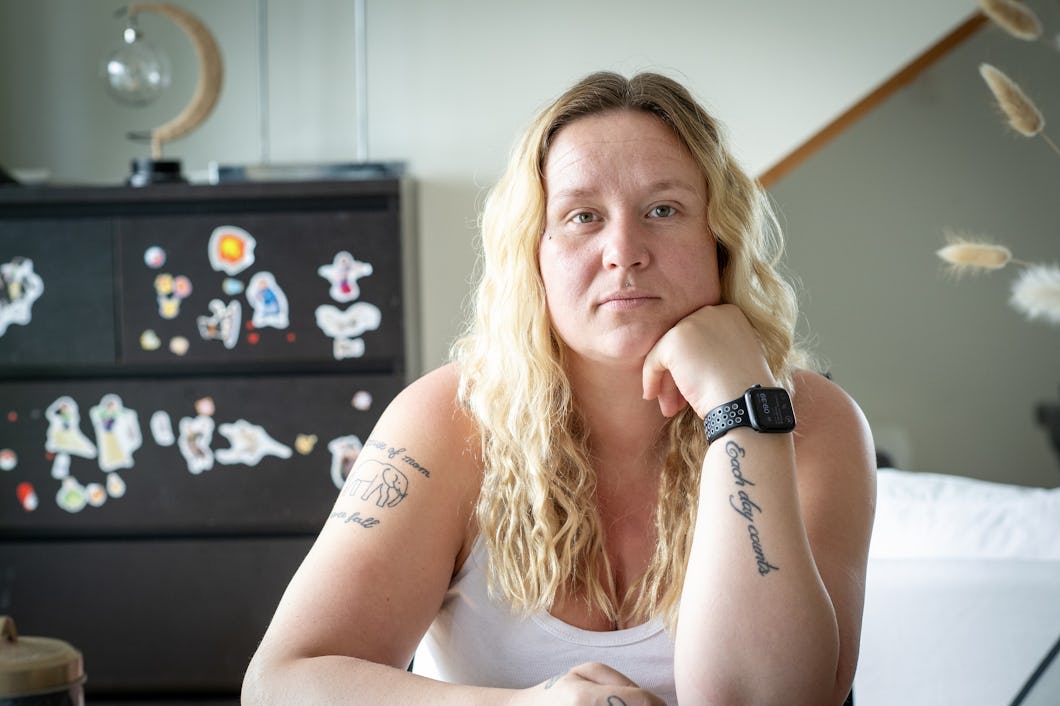 Camilla Dahlgren med långt blont hår sitter inomhus och stödjer hakan mot handen. Hon har synliga tatueringar på båda armarna och bär en smartklocka. I bakgrunden finns en byrå med klistermärken.