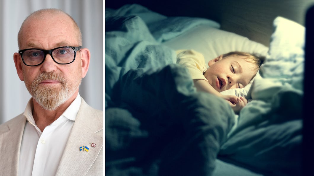 Johan Lindholm med glasögon och kostym står framför kameran. Bredvid honom sover ett barn i en säng under en filt.