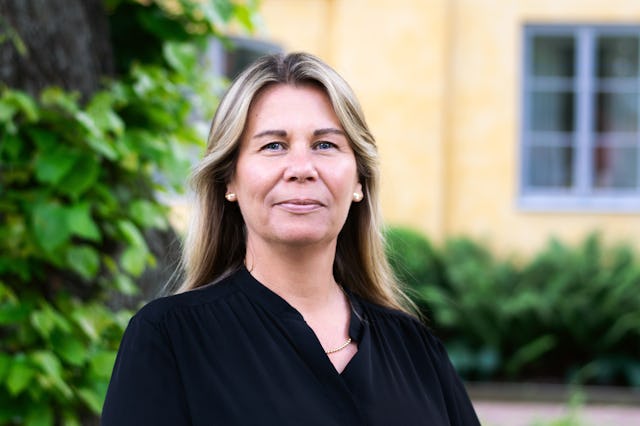 En kvinna med långt blont hår och en svart topp står utomhus utomhus och gul byggnad med växtlighet i bakgrunden.