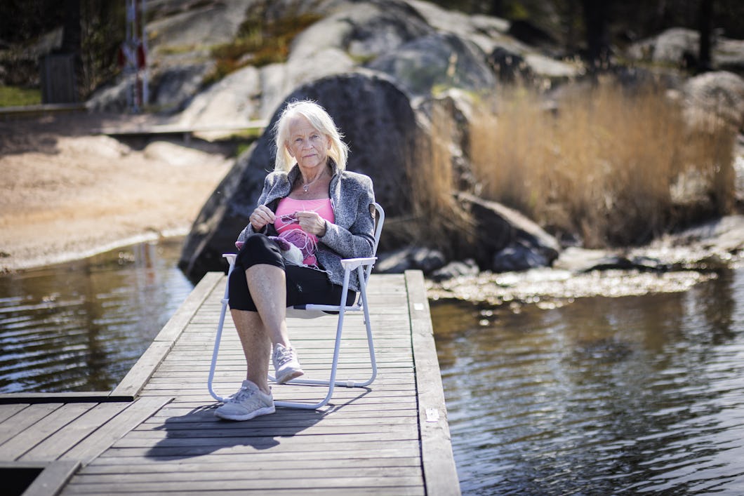 En kvinna sitter på en fällstol vid slutet av en brygga över vatten. Hon håller i stickor. I bakgrunden finns stenar och torrt gräs.