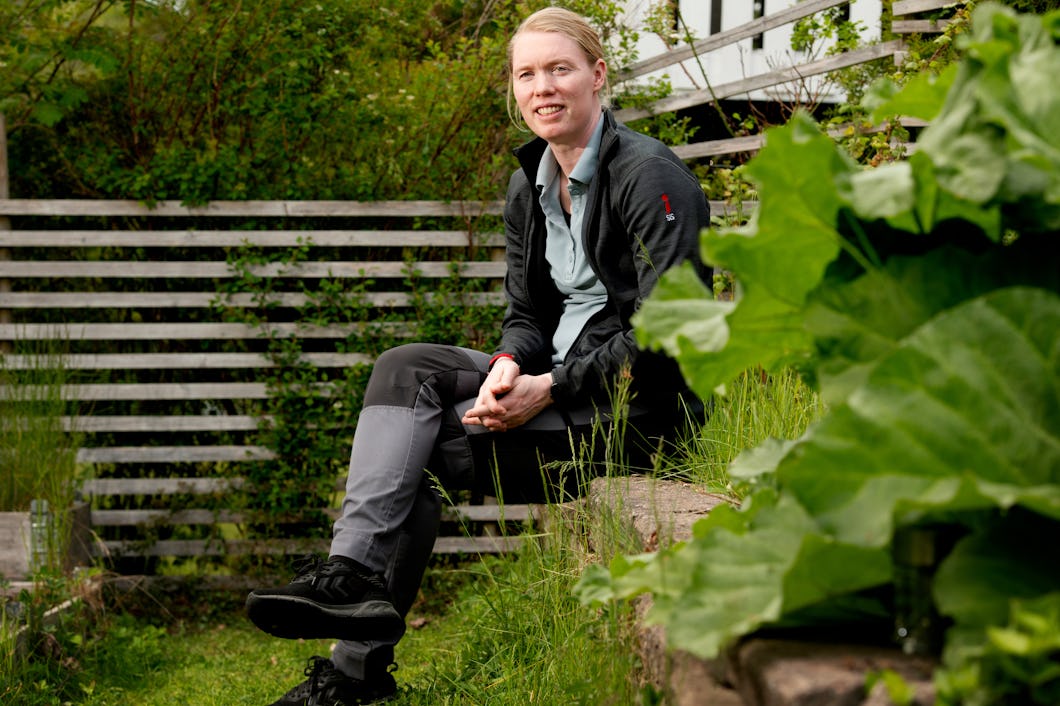 Hedvig Lindahl sitter på en stenmur i en trädgård. Runtom finns växter och i bakgrunden syns ett staket av trä.