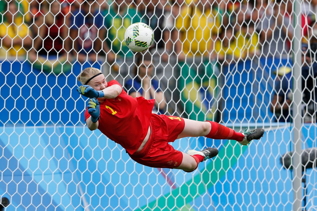 Hedvig Lindahl kastar sig åt vänster i luften för att stoppa en boll nära målet, medan publik syn i bakgrunden.