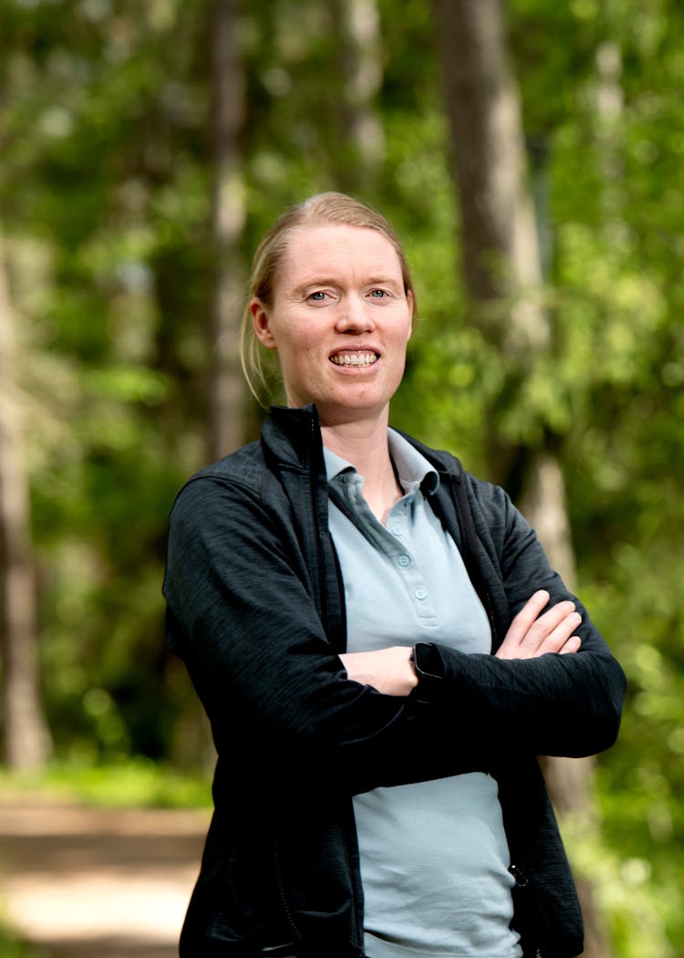 Hedvig Lindahl står på en skogsstig med armarna i kors, iklädd en blå skjorta och svart jacka. I bakgrunden syns träd och växtlighet.