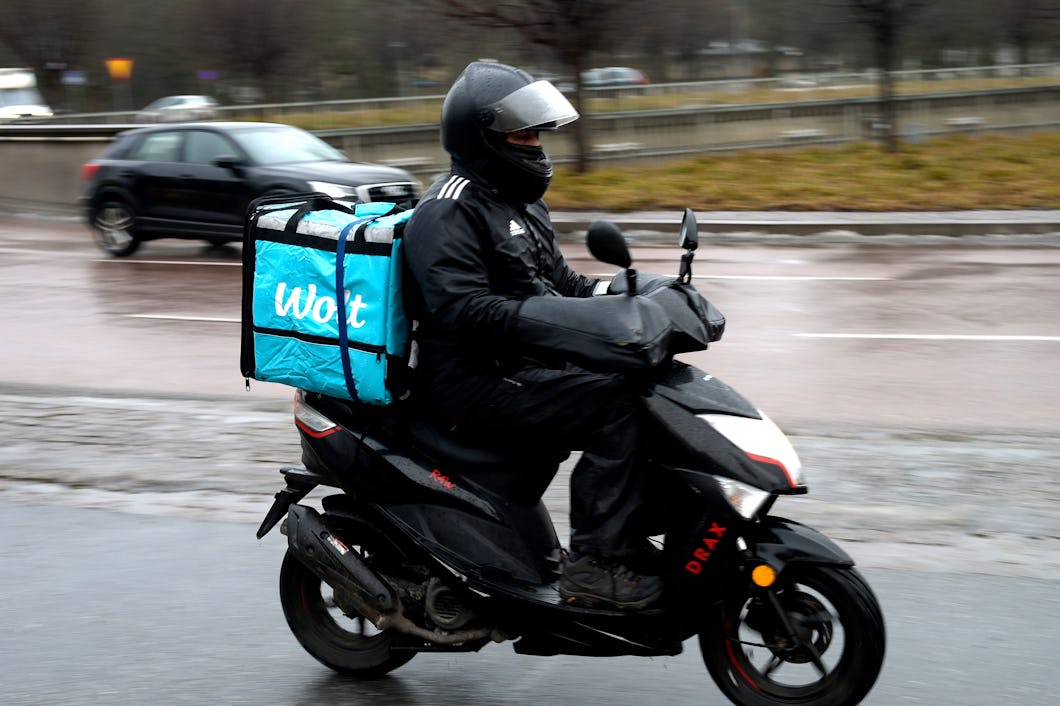 En matbud kör en scooter på en våt stadsgata och har regnkläder samt en Wolt-väska på sig.