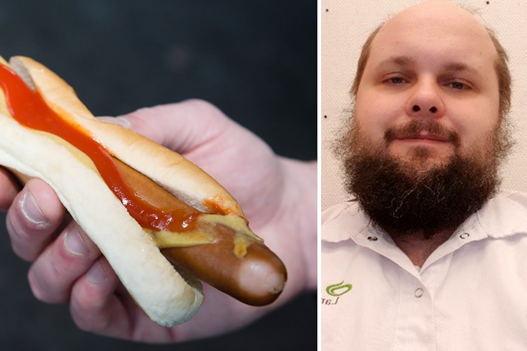 En person håller i en korv med ketchup och senap. Bredvid står en man med skägg.