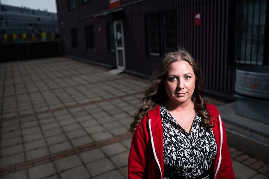 Carolina Hjortzberg med långt brunt hår och en röd jacka står på ett asfalterat område utanför en byggnad och tittar in i kameran.