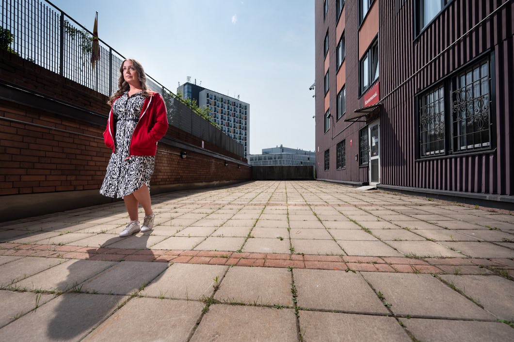 Carolina Hjortzberg med röd jacka och mönstrad klänning står på en stenlagd gård bredvid en flervåningsbyggnad under dagen.