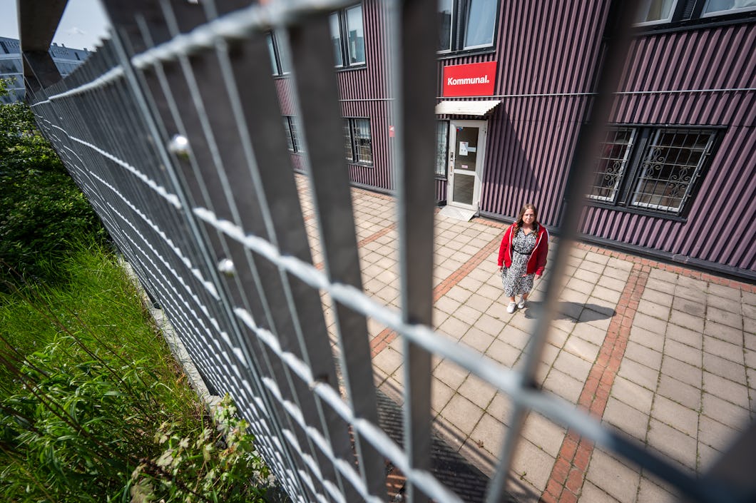Carolina Hjortzberg i röd tröja står framför en byggnad med skylten ”Kommunal”, sedd genom ett staket av metall.