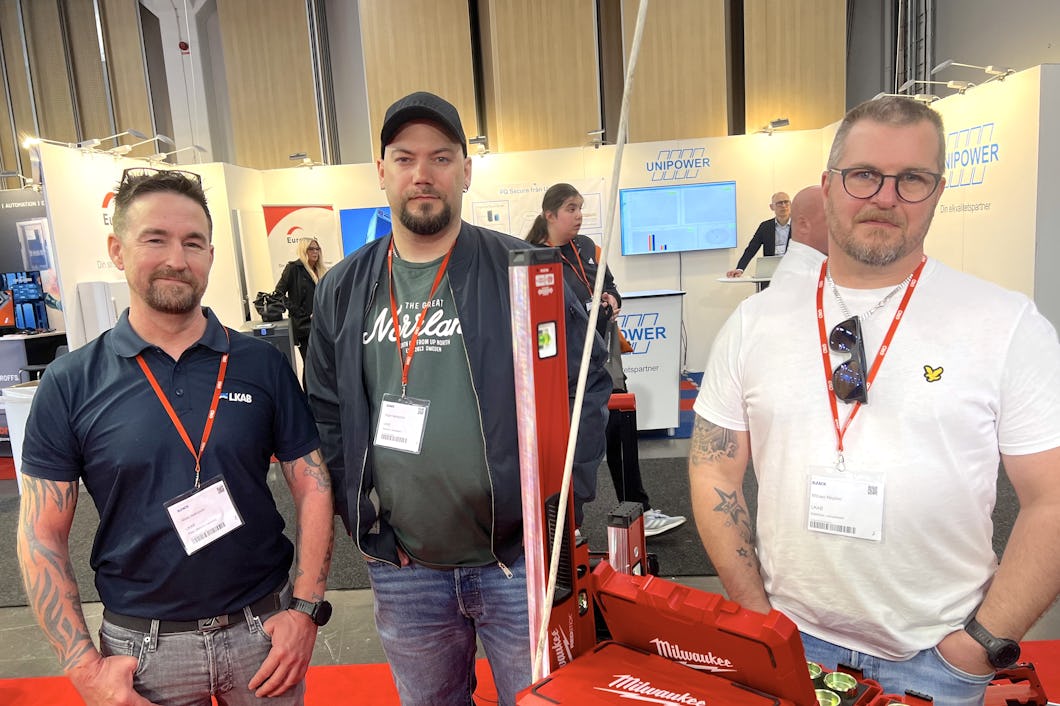 Tre män står inomhus vid en monter. Alla har namnskyltar och röda band runt halsen. Framför dem finns utrustning och verktyg med Milwaukee-logotypen.