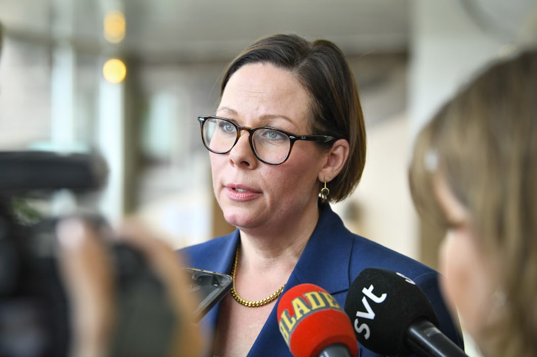 En kvinna med glasögon och blå kavaj talar till media medan flera mikrofoner hålls upp mot henne.