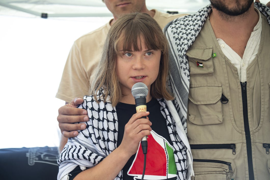 En kvinna talar i en mikrofon. Hon har en svartvit palestinasjal och en tröja med en vattenmelon på sig. Två personer står bredvid henne.