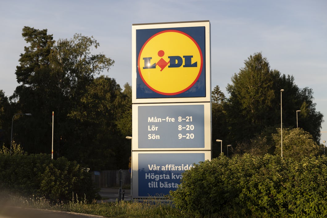 En skylt för Lidl visar butikens öppettider för vardagar, lördagar och söndagar, med träd och växtlighet i bakgrunden.