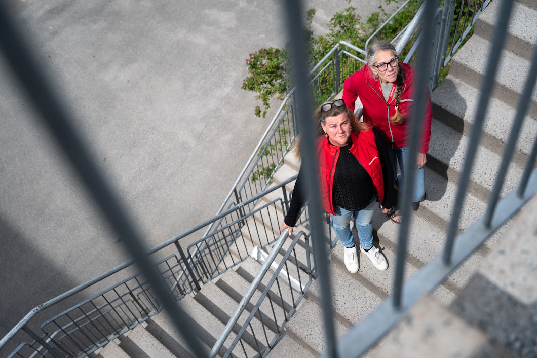 Annie Lisstrand och Cecilia Holm i röda jackor står på en utomhustrappa av betong och tittar upp mot kameran genom metallräcken.