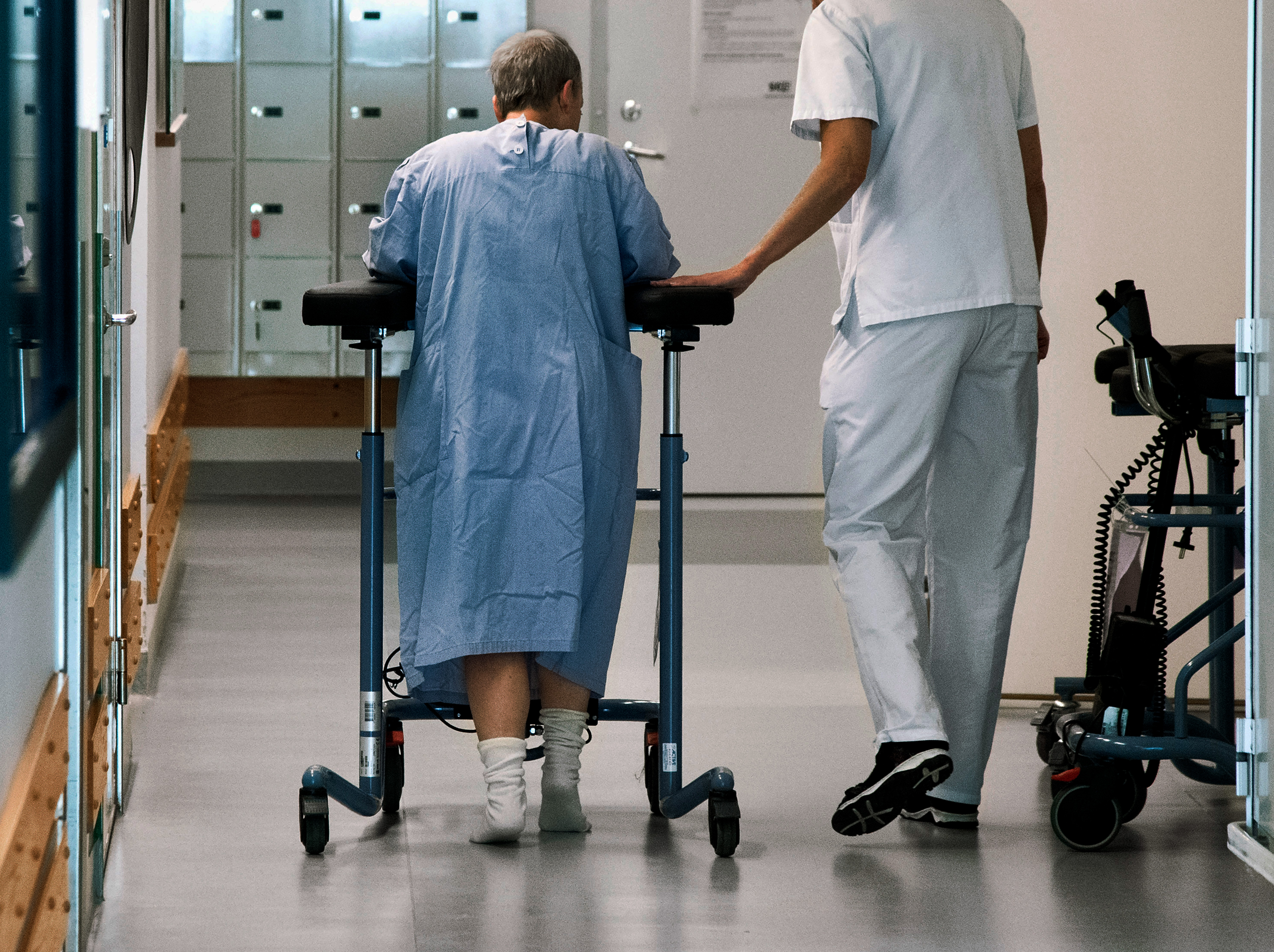En äldre patient bär sjukhuskläder och går med hjälp av en rullator, assisterad av en vårdpersonal i vit uniform, längs en korridor.